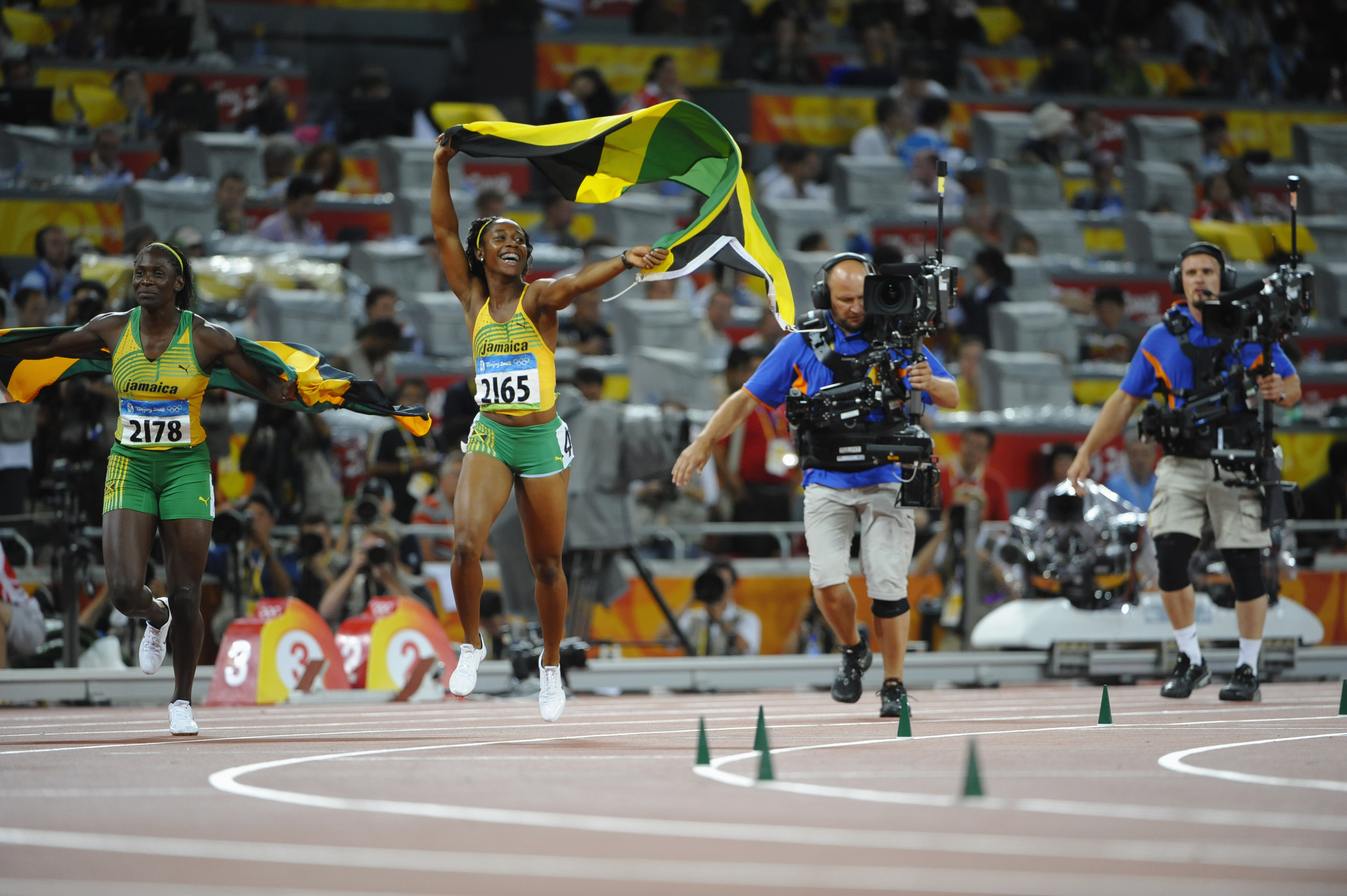 JO Beijing 2008, Athlétisme, 100m Femmes - Finale, Kerron STEWART (JAM) 2e et Shelly-Ann FRASER (JAM) 1e font un tour de piste avec le drapeau jamaicain, suivies par des cameramen.